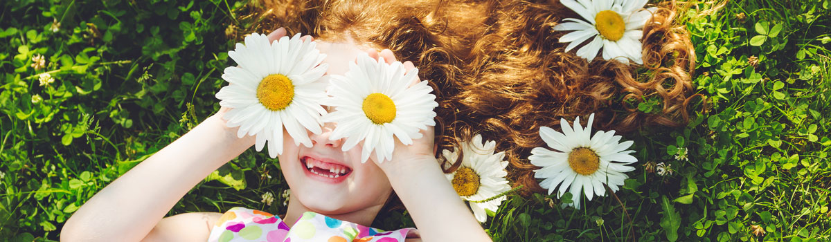young girl laughing while lying in grass while covering her eyes with white daisies Colorado Allergy & Asthma Centers
