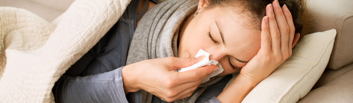 woman using tissue on her nose while in bed with the flu Colorado Allergy & Asthma Centers